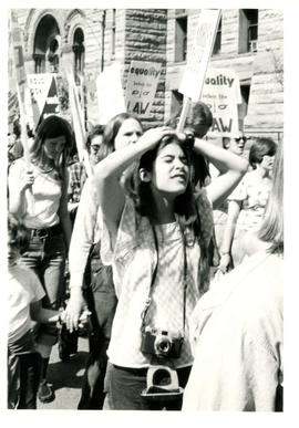 Woman demonstrator marching with arms on top of head holding camera surrounded with some people h...