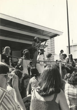 Unidentified woman on a stage outdoors, speaking to a group of women