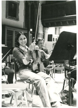 Seated orchestra members holding their violins during a poetry reading's rehearsal break, [Guelph]