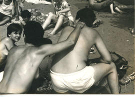 Woman with one hand on shoulder of woman sitting on blankets at the beach with their back to the ...