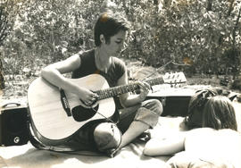 Unidentified woman sitting on a blanket on the ground and playing the guitar