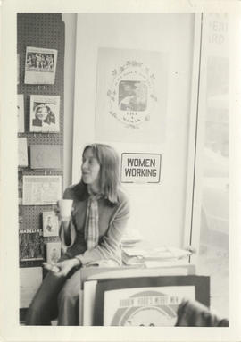 Unidentified woman clutching a styrofoam cup and sitting on a desk in [A Woman's Place, Toronto]